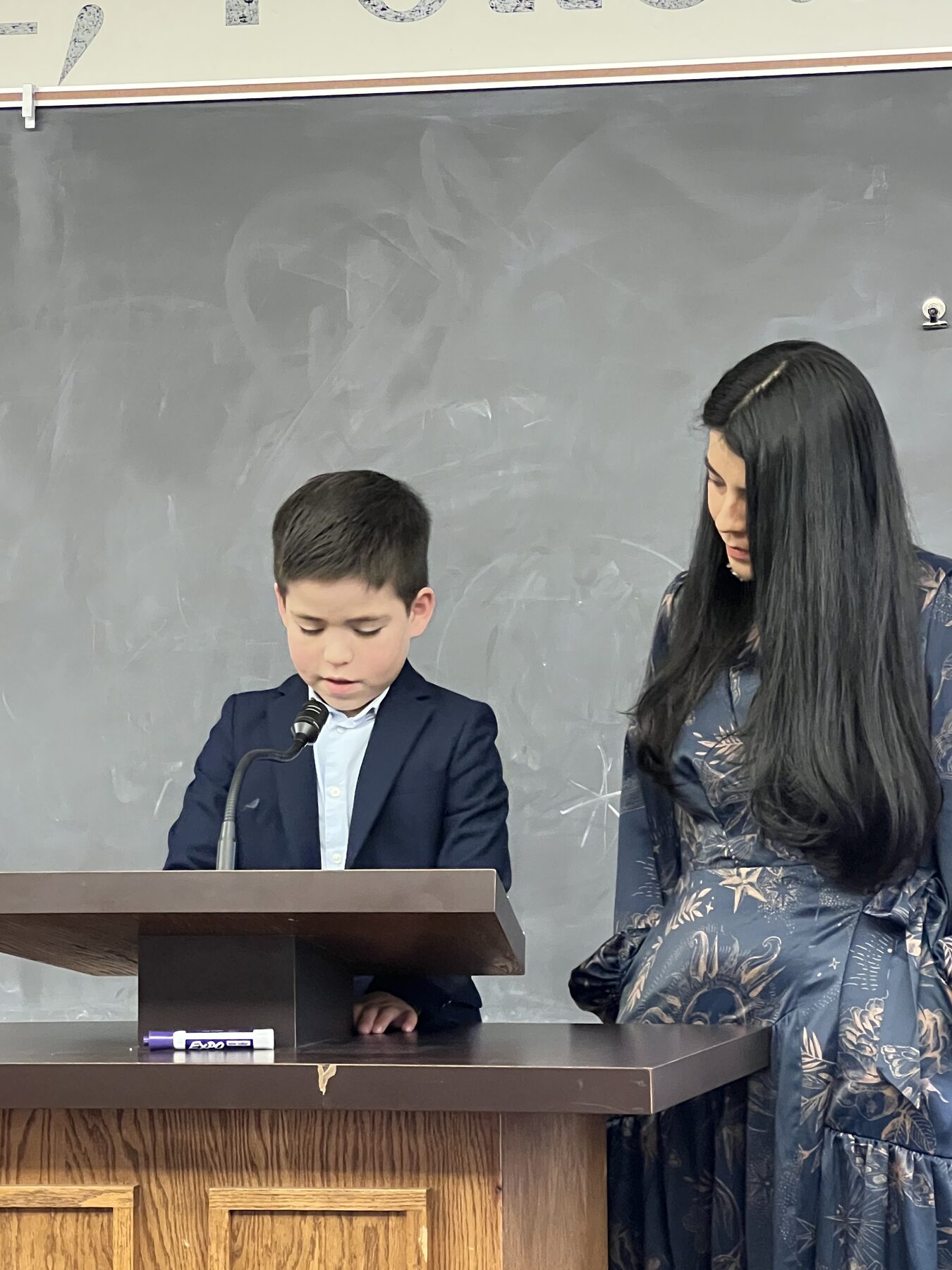 Oliver speaking at a church classroom podium beside his mom.